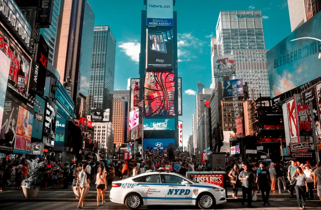 Crowds in Times Square, New York City, surrounded by bright billboards and an NYPD patrol car in the foreground.