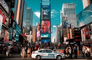 Crowds in Times Square, New York City, surrounded by bright billboards and an NYPD patrol car in the foreground.